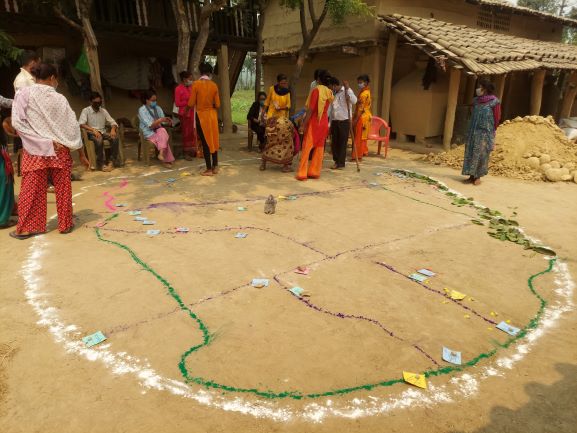 A group of people gathered around a large circular ground drawing, likely for an outdoor activity or event, in a rural setting with some seated participants and scattered materials.