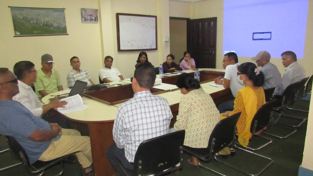 A group of people are sitting around a large conference table in a room, engaged in a meeting. A blank screen and a whiteboard are visible on the walls.