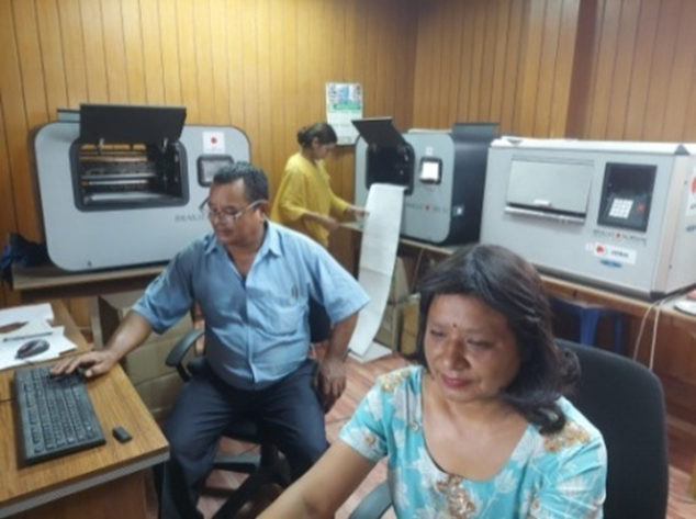 one man and women working in computer. behind them are braille printers.